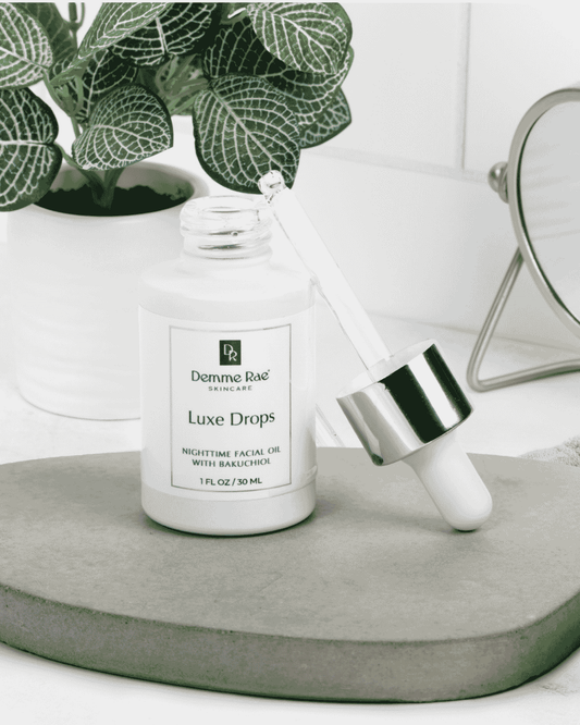 Demme Rae Skincare Luxe Drops in a white bottle with a white dropper and silver collar, styled on a gray stone tray with a potted plant and mirror in the background.
